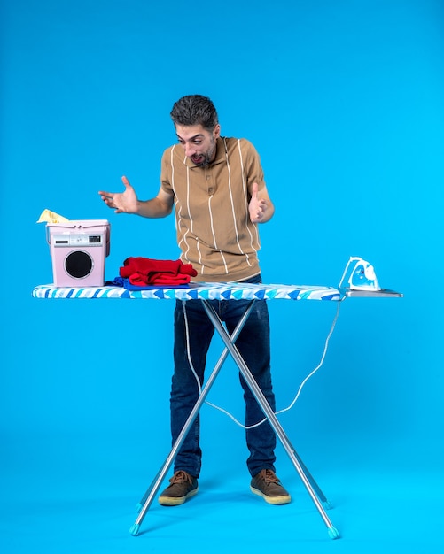 Vue de face jeune homme derrière une planche à repasser sur fond bleu couleur les travaux ménagers blanchisserie homme émotion propre machine à laver