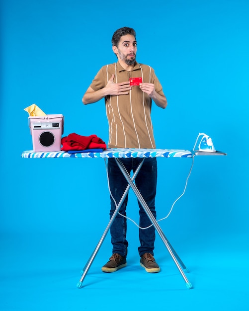 Vue de face jeune homme derrière une planche à repasser tenant une carte bancaire rouge sur fond bleu argent blanchisserie lave-linge couleur fer travaux ménagers