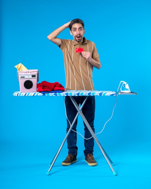 Vue de face jeune homme derrière une planche à repasser tenant une carte bancaire rouge sur fond bleu argent laver le linge ménage machine à laver fer à repasser