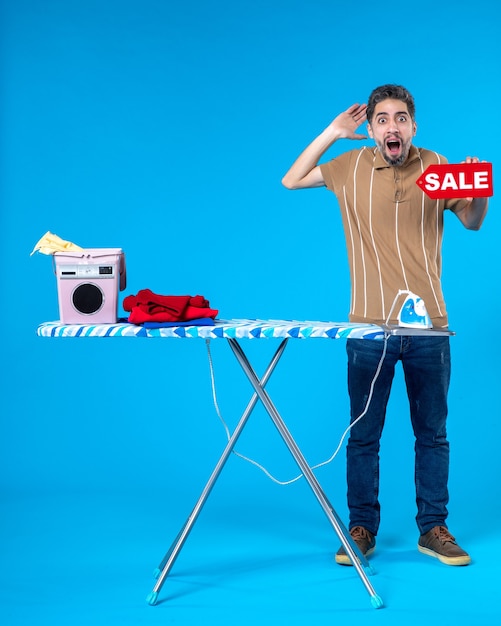 Vue de face jeune homme derrière une planche à repasser tenant une vente rouge écrit sur fond bleu shopping femme au foyer machine à laver blanchisserie ménage