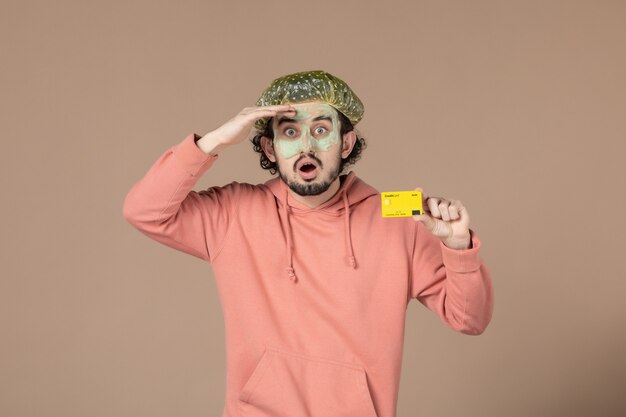 vue de face jeune homme avec masque sur son visage tenant une carte de crédit sur fond marron argent thérapie salon de soins de la peau du visage soins du corps spa