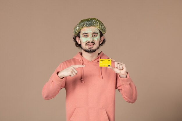 vue de face jeune homme avec masque sur son visage tenant une carte de crédit sur fond marron salon de thérapie capillaire peau du visage soins du corps spa