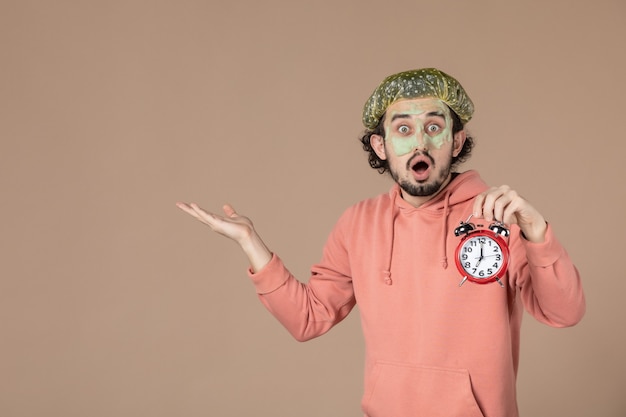 Photo vue de face jeune homme avec masque tenant des horloges sur fond marron temps de thérapie spa salon peau soins du corps couleur du visage