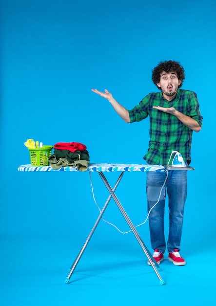 Vue de face jeune homme avec planche à repasser sur fond bleu maison lave-linge travaux ménagers couleur humaine propre