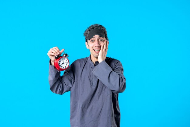 Vue de face jeune homme en pyjama tenant des horloges rouges sur fond bleu cauchemar sombre insomnie couleur sommeil sommeil fin de nuit lit de rêve