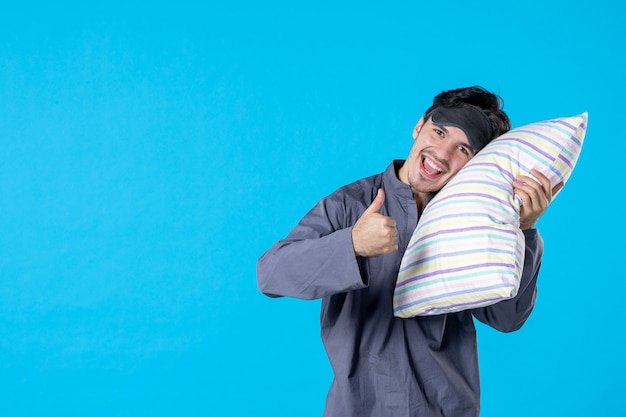 Vue de face jeune homme en pyjama tenant un oreiller sur fond bleu service lit repos couleur rêve sommeil nuit humaine cauchemar tardif