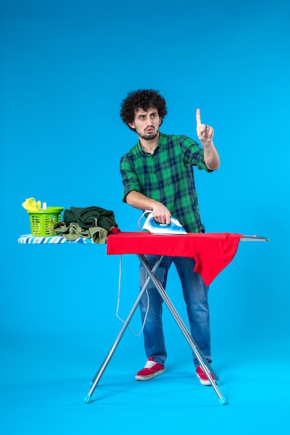 Vue de face jeune homme repassage des vêtements rouges à bord sur fond bleu maison de couleur machine à laver les travaux ménagers de l'homme