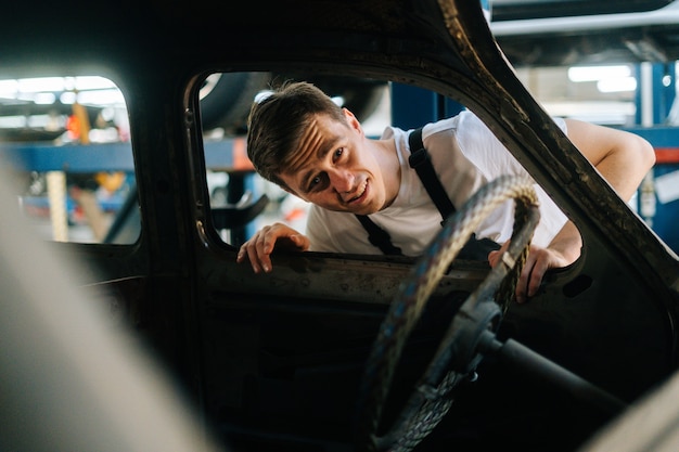 Vue de face d'un jeune homme de service frustré en uniforme inspectant l'intérieur d'une vieille voiture en réparation automobile