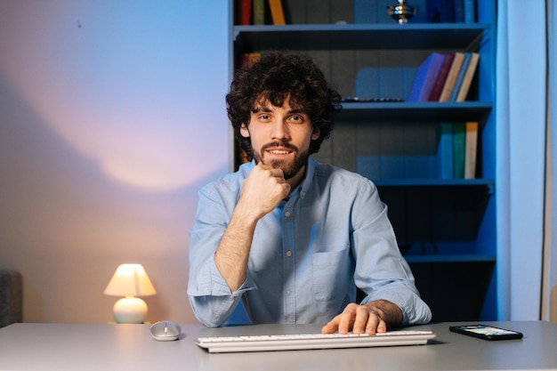 Vue de face d'un jeune homme souriant tapant un message en ligne sur un clavier sans fil en regardant la caméra