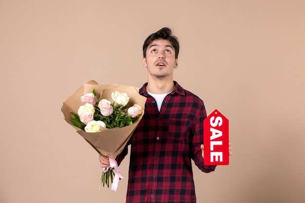 Vue de face jeune homme tenant de belles fleurs pour femme et plaque signalétique de vente sur mur marron