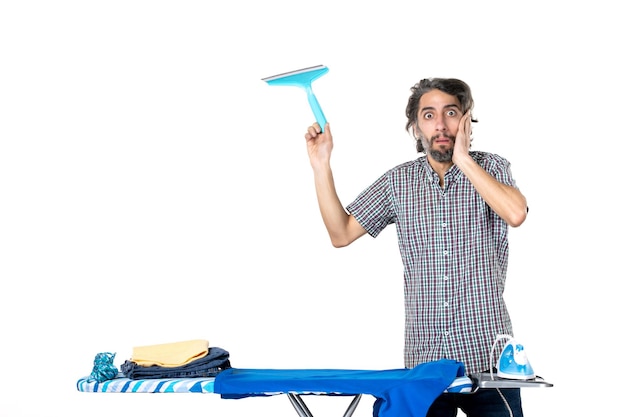 vue de face jeune homme tenant une brosse plus propre derrière une planche à repasser surpris sur un fond clair accueil fer à repasser linge vêtements machine propre travaux ménagers