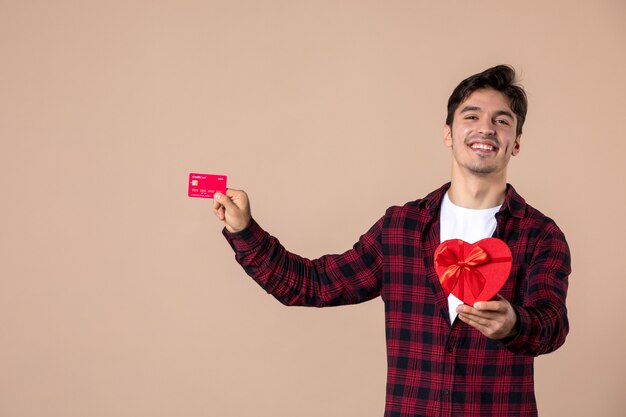 Vue de face jeune homme tenant un cadeau en forme de coeur et une carte bancaire sur un mur marron