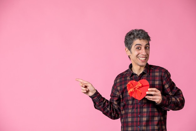 Vue de face jeune homme tenant un cadeau en forme de coeur rouge sur un mur rose