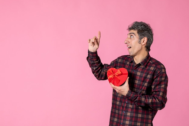 Vue de face jeune homme tenant un cadeau en forme de coeur rouge sur un mur rose