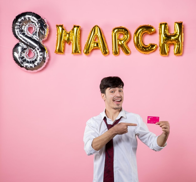 Vue de face jeune homme tenant une carte bancaire rose avec décoration de mars sur fond rose fête présente vacances homme féminin argent égalité couleurs shopping