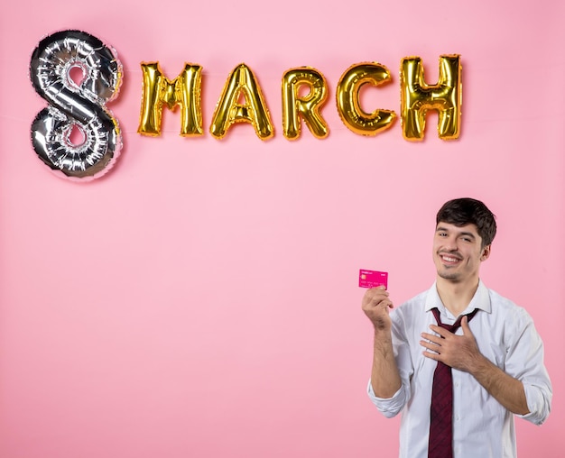 Vue de face jeune homme tenant une carte bancaire rose avec décoration de mars sur fond rose présente l'égalité féminine homme de vacances shopping couleur argent