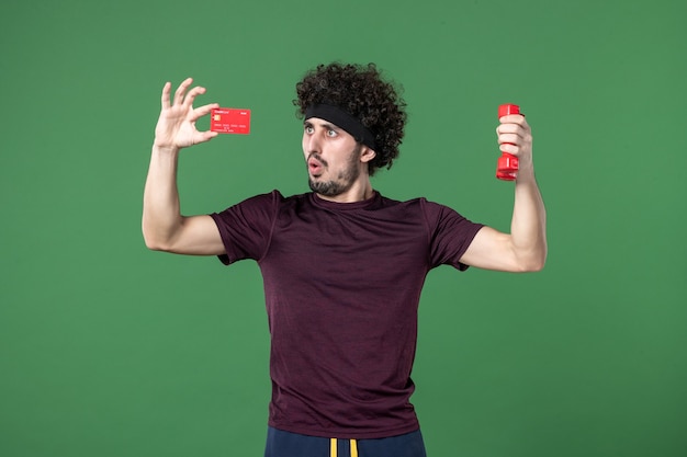 Vue de face jeune homme tenant des haltères et une carte bancaire sur fond vert athlète d'entraînement sport modèle shopping couleur gym