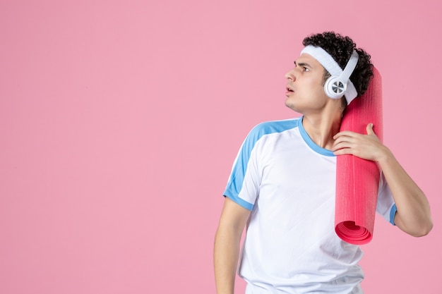 Vue de face jeune homme en vêtements de sport avec tapis de yoga sur mur rose