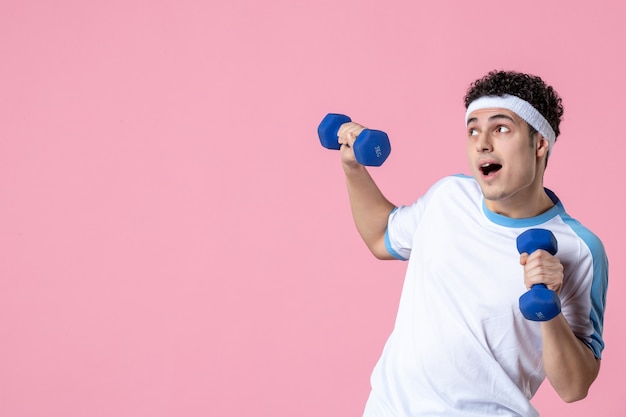 Vue de face jeune homme en vêtements de sport travaillant avec des haltères sur un mur rose