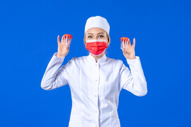 vue de face jeune infirmière en costume médical avec masque rouge tenant des flacons sur fond bleu virus de la santé covid- hôpital médicament médecin médecine couleurs pandémiques