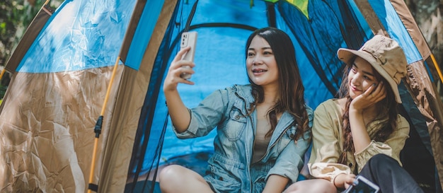 Vue de face Jeune jolie femme asiatique et sa petite amie assise devant la tente utilisent un téléphone portable pour prendre des photos pendant le camping en forêt avec bonheur ensemble
