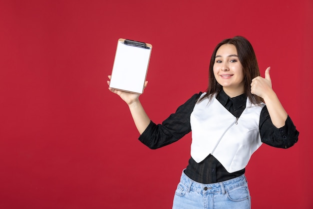 vue de face jeune jolie serveuse en uniforme tenant un petit bloc-notes pour les commandes sur fond rouge travail beauté femme emploi travailleur dîner soirée