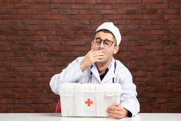 Vue de face jeune médecin bâillant en costume médical blanc avec trousse de premiers soins sur mur de briques marron