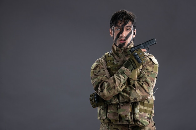 Vue de face jeune soldat combattant en tenue de camouflage avec arme à feu sur le mur sombre