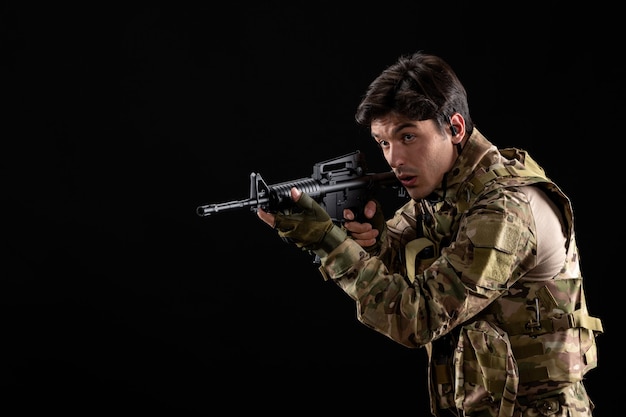 Vue de face jeune soldat en uniforme visant son fusil sur mur noir