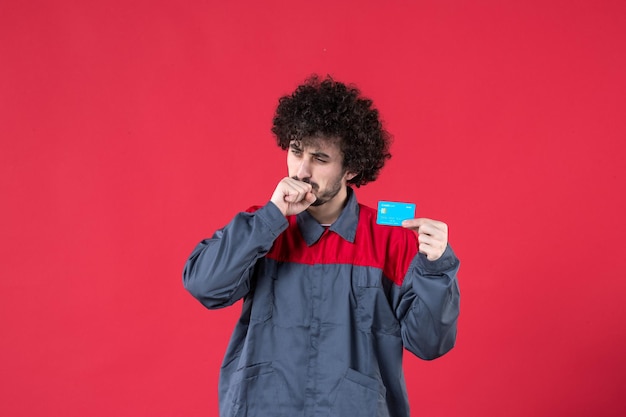Vue de face jeune travailleur masculin en uniforme tenant une carte bancaire bleue sur une surface rouge