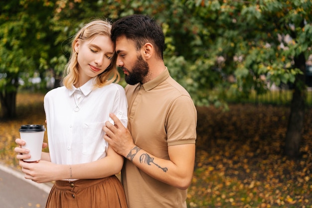 Vue de face d'un joli jeune couple amoureux debout embrassant les yeux fermés au parc de la ville sur fond d'arbres Homme barbu et charmante femme marchant