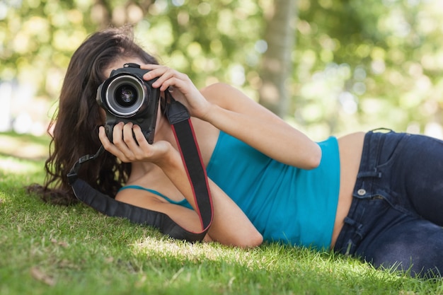 Vue de face de la jolie femme brune allongée sur une pelouse prenant une photo