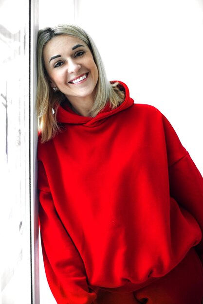Vue de face d'une jolie jeune femme blanche souriante en sweat à capuche rouge et pantalon debout dans une salle blanche à l'air heureux