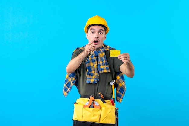 Vue de face mâle constructeur en casque jaune tenant une carte bancaire sur fond bleu argent architecture bâtiment constructeur travailleur couleurs plates