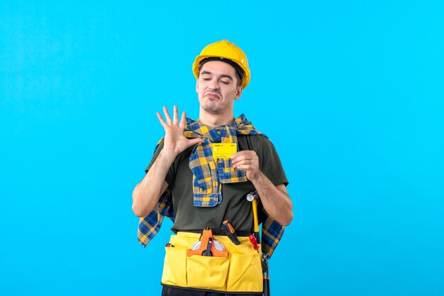 Vue de face mâle constructeur en casque jaune tenant une carte bancaire sur fond bleu couleur argent architecture plate constructeurs ouvriers du bâtiment