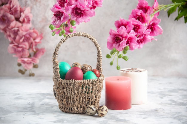 vue de face oeufs de pâques colorés à l'intérieur d'un panier conçu avec des fleurs et des bougies sur une surface blanche