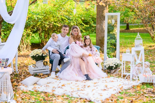 Vue de face de parents et d'enfants élégants souriant et regardant la caméra assis sur un canapé confortable dans un parc décoré par un beau décor et des fleurs en pot pendant la journée d'automne