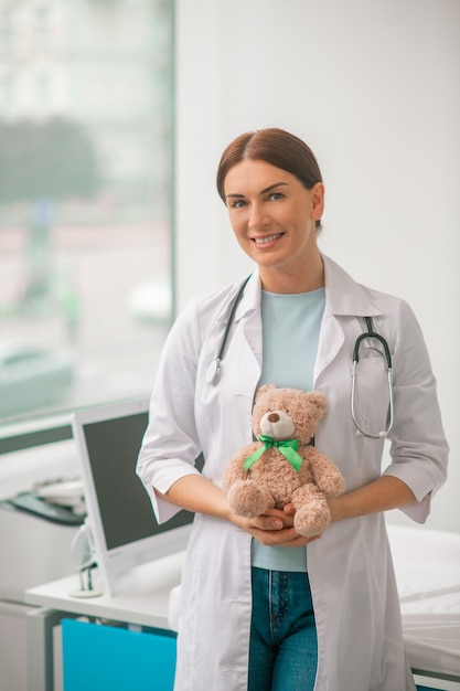 Vue de face d'un pédiatre aux cheveux noirs satisfait avec un stéthoscope et un ours en peluche regardant vers l'avenir