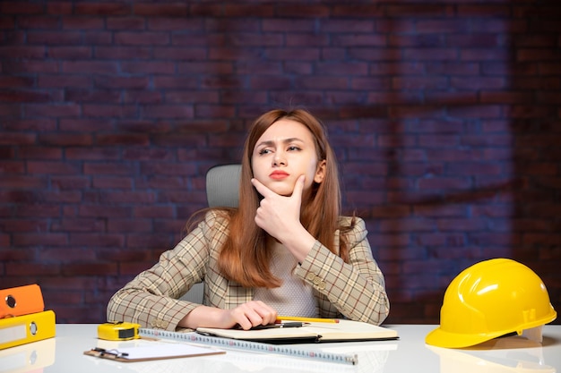 Photo vue de face pensant travailleuse dans le chantier de construction assis derrière la table de l'ordre du jour de l'emploi ingénieur chef d'entreprise plan d'entreprise de travail