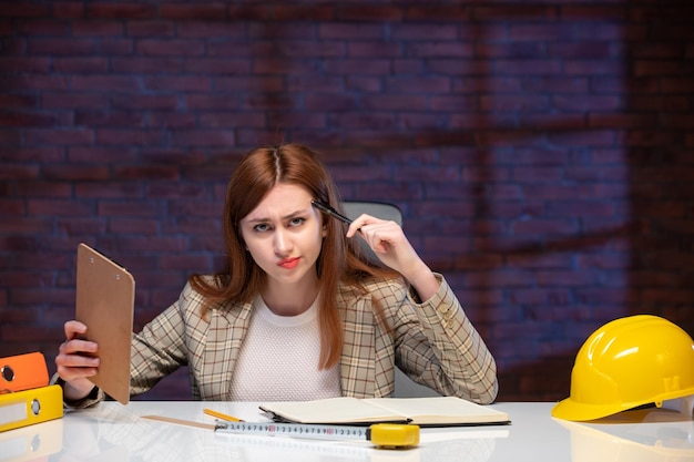 Vue de face pensant une travailleuse à l'intérieur du chantier de construction assis derrière une table avec des documents d'entreprise entrepreneur chef de projet plan de projet ingénieur