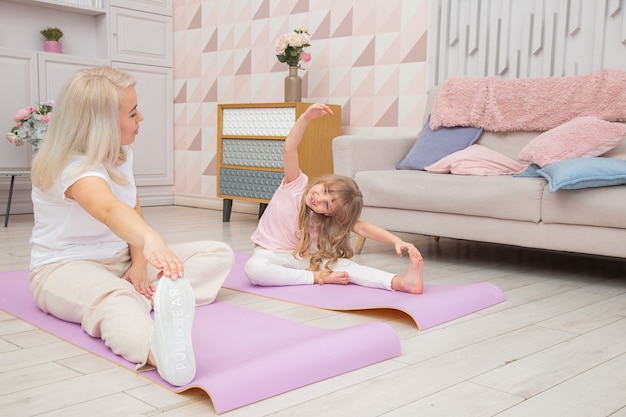 Vue de face pleine longueur mère blonde souriante sur tapis de yoga avec jolie petite fille d'âge préscolaire ludique faire divers exercices. Heureuse maman pratiquant le yoga avec un petit enfant.
