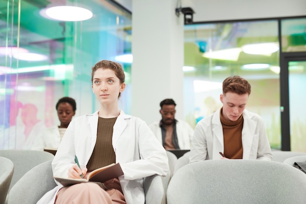 Vue de face portrait d'une jeune femme portant une blouse de laboratoire assise dans le public et écoutant une conférence sur la médecine à l'université ou dans un centre de coworking, espace de copie