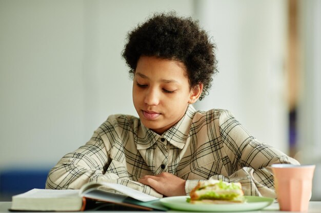 Vue de face portrait d'une jeune fille afro-américaine lisant un livre pendant la pause déjeuner à la cantine scolaire