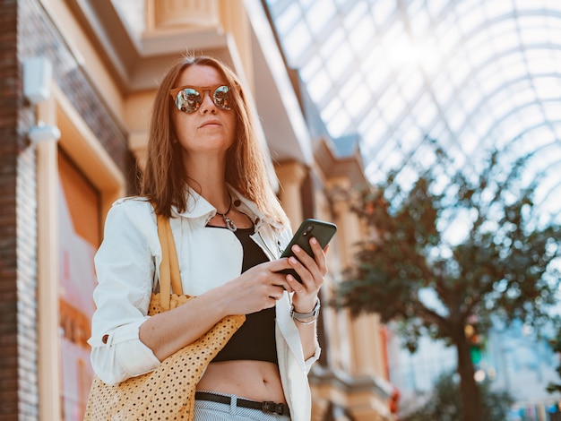 Vue de face portrait mode moderne femme heureuse hipster marchant et utilisant un téléphone intelligent dans une rue de la ville portant des lunettes de soleil.