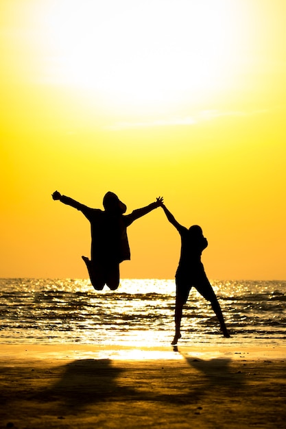 Vue de face de silhouette jeune homme et jeune fille sautant par-dessus la plage de la mer avec fond de coucher de soleil.