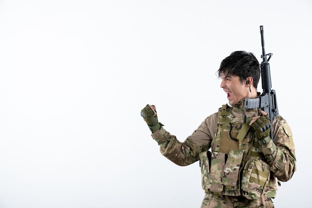 Vue de face d'un soldat masculin heureux avec une mitrailleuse en camouflage sur un mur blanc