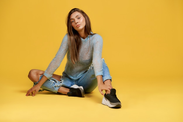 Vue de face de la terre de dame confiante touchant la jambe. Belle jeune femme portant des jeans déchirés. Isolé sur un mur jaune avec espace copie. Concept de beauté et de confiance.