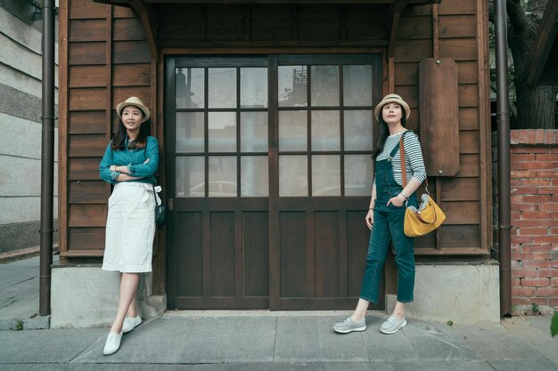 vue de face sur toute la longueur de deux voyageurs asiatiques debout près de la porte en bois d'une maison ancienne japonaise traditionnelle. charmantes femmes touristes s'appuyant sur un mur en bois avec espace de copie style vintage extérieur.