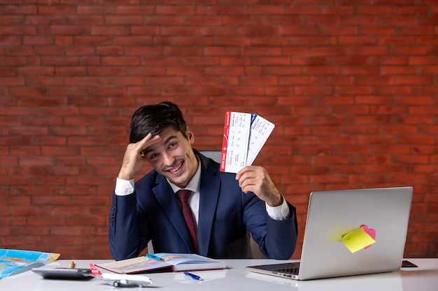 Vue de face d'un travailleur masculin assis derrière son lieu de travail en costume tenant une paire de billets d'avion travail à l'étranger projet de tourisme d'emploi occupation d'entreprise