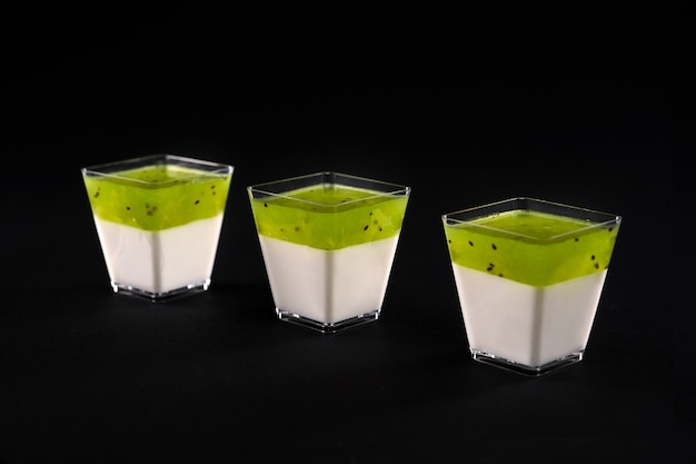 Vue De Face De Trois Petits Verres Carrés Avec Panna Cotta Laiteuse. Délicieux Dessert Sucré Décoré De Garniture De Pomme Vert Vif, Isolé Sur Fond Noir. Concept Alimentaire.